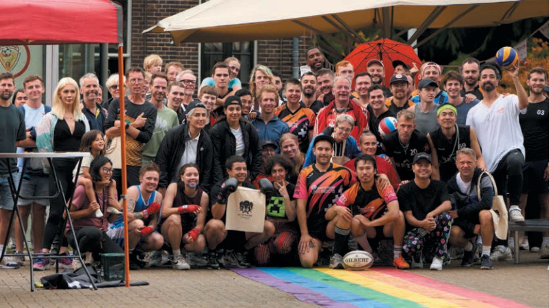 Groepsfoto van divers sportteam op regenboogkleuren, met rugbybal, volleybal en bokshandschoenen. Inclusie en sportiviteit benadrukt.