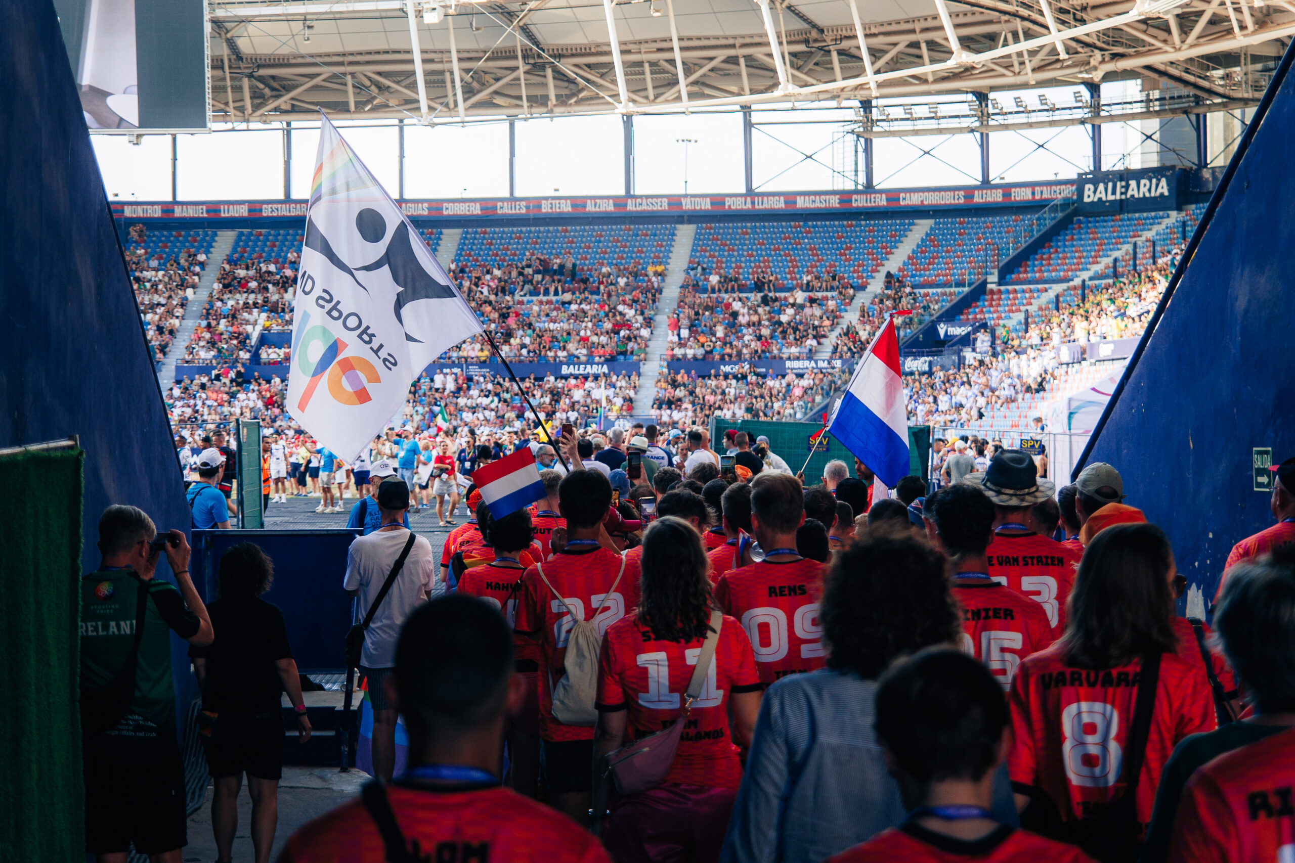 Deelnemers in rode shirts lopen een stadion binnen, begeleid door vlaggen waaronder een vlag met het logo van AD Sports.
