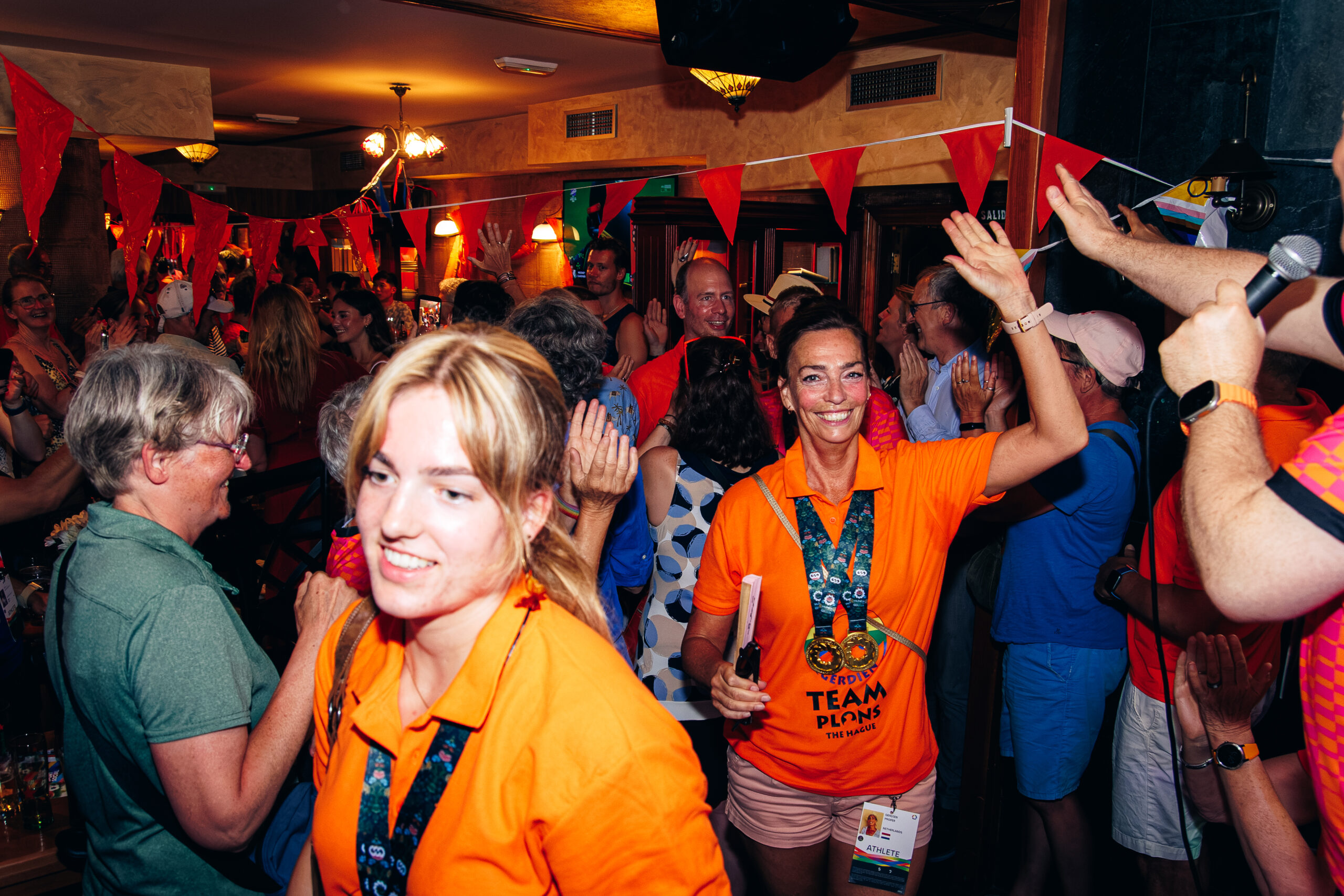 Een glimlachende vrouw in een oranje shirt met twee medailles zwaait naar een feestende menigte in een zaal versierd met rode vlaggetjes.