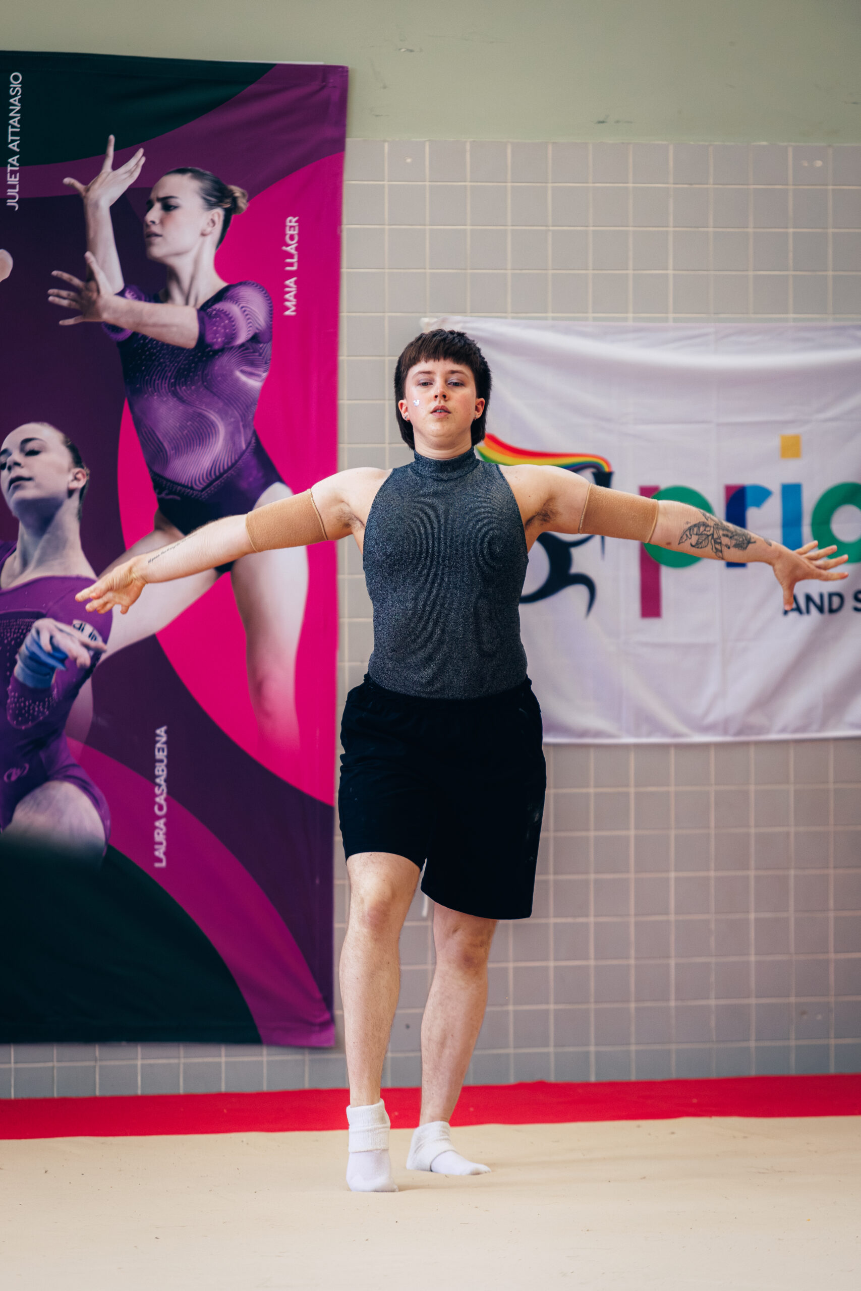 Persoon met kort haar in sportkleding staat in een turnhouding met gespreide armen voor een spandoek met gymnasten en een pride-vlag.