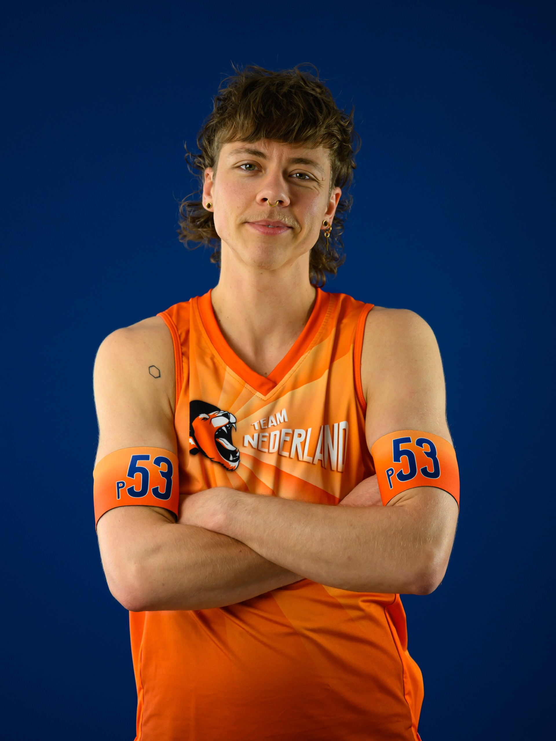 Roller derby speler van Team Nederland, nummer 53, in oranje shirt met armen gekruist op blauwe achtergrond.