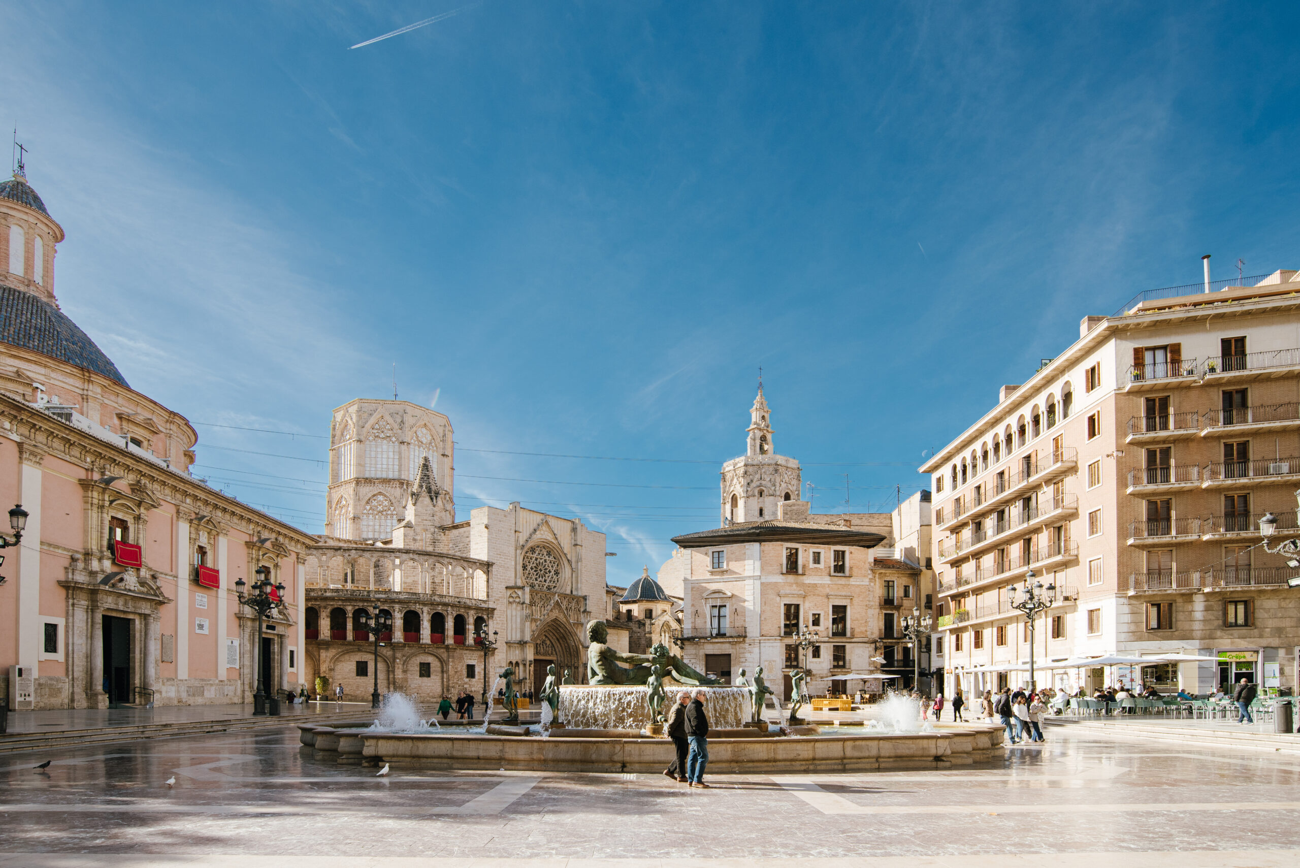 plazavirgen_valencia (10) Panoramic view of Plaza de la Virgen in Valencia, Spain, featuring the central fountain, historic buildings including the Valencia Cathedral and Basilica, under a clear blue sky.