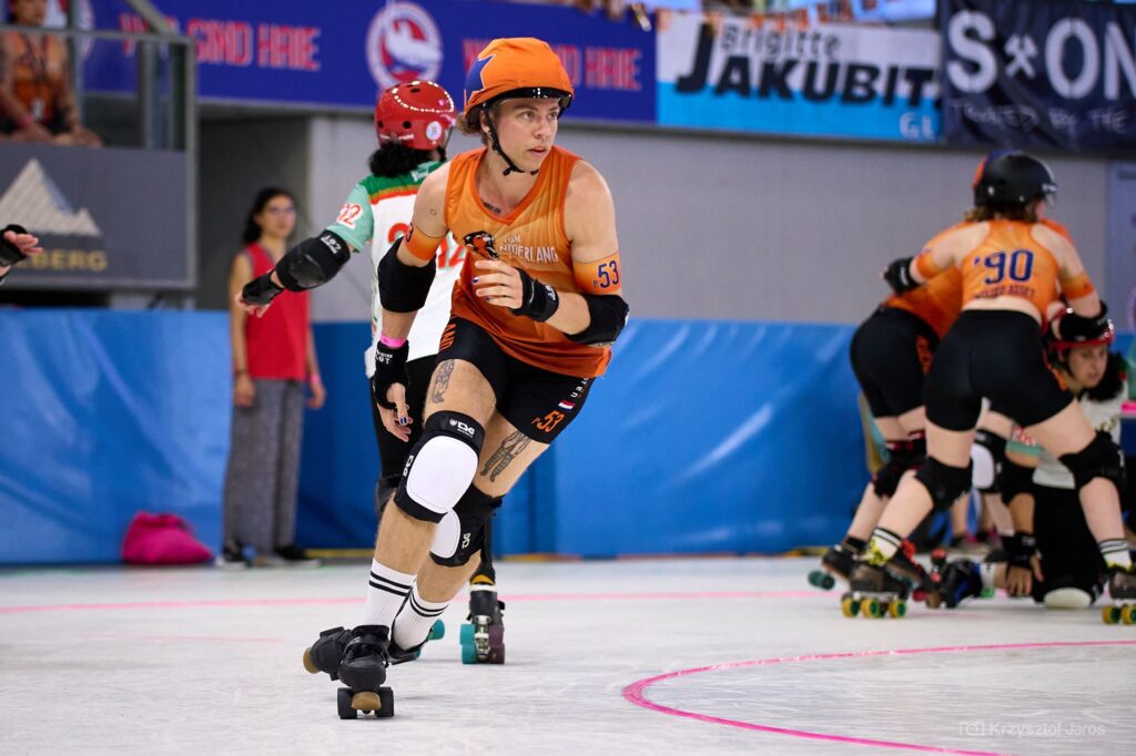 Veer Vesper Visser, roller derby-speler in actie. Oranje tenue, nummer 53. Snelle beweging op de baan. Spectaculaire sportfoto. Roller derby kampioenschap.